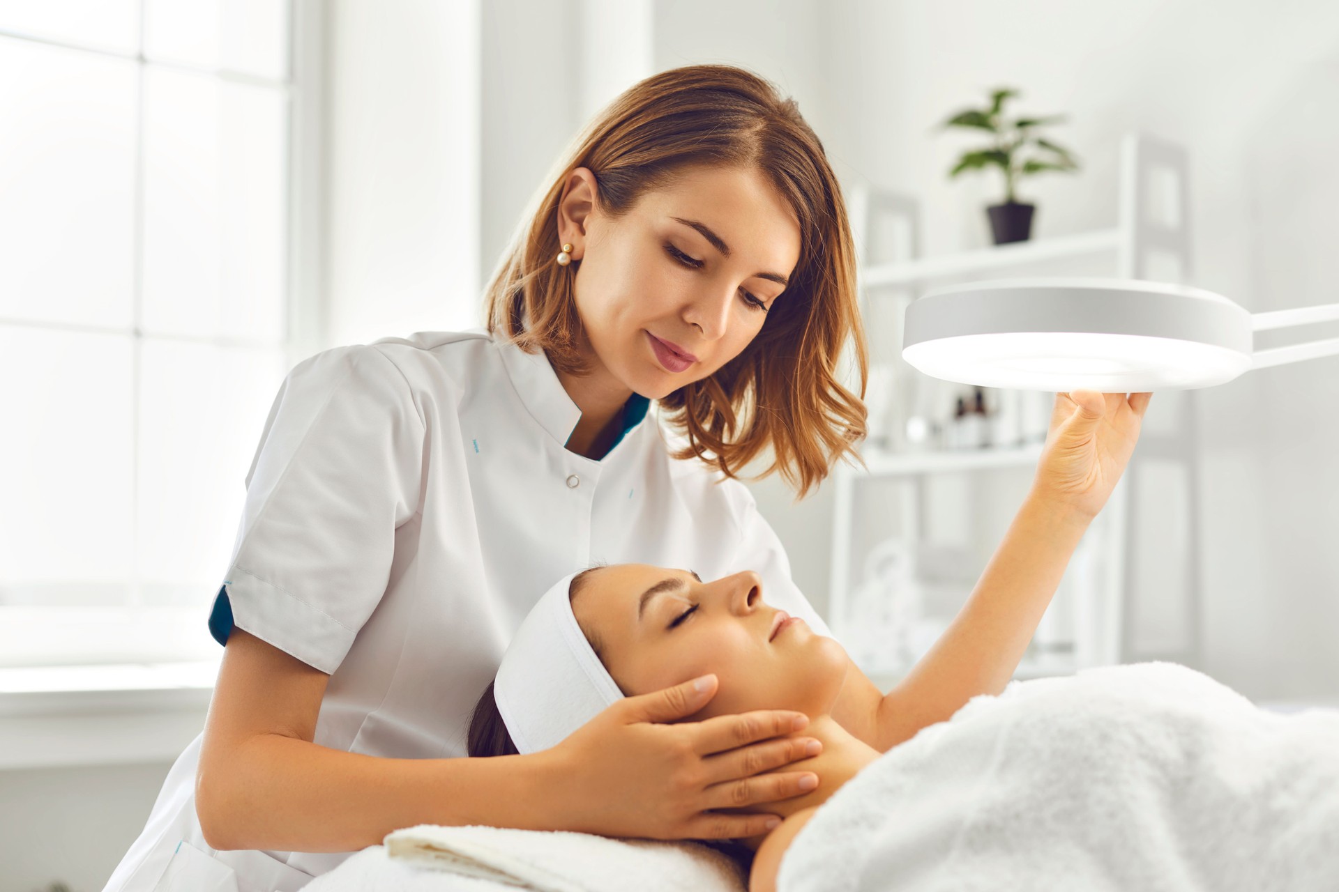Cosmetologist directing lamp to woman for facial treatment
